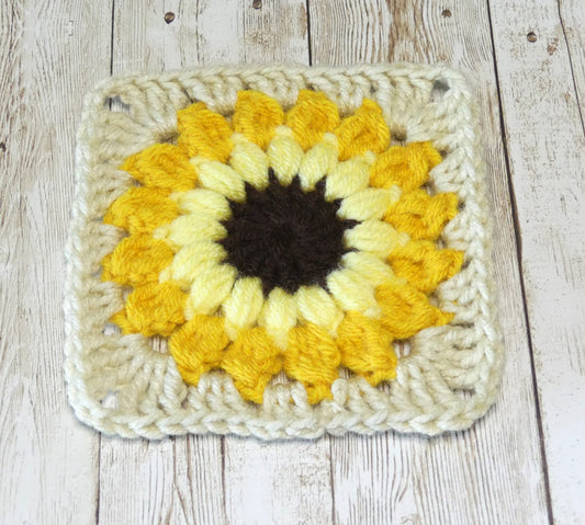 A crocheted granny square that looks like a sunflower with a brown center, light yellow 2nd row, gold 3rd row and cream colored 4th row